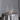 The Hall of Flame Round Table Cluster features a round table with a silver satin tablecloth, gold flatware for four, empty coupe glasses, a white floral centerpiece, and seven long-burning tall white candles in silver holders.