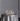 A Hall of Flame Round Table stands out in the room, elegantly covered with a silver satin cloth, gold flatware, two wine glasses, white flowers centerpiece, and Pillar Cluster candles from Hall of Flame, set against a plain gray wall.