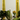 The Hall of Flame Olive Green Wax Candle Holder features a pillar candle, surrounded by leafy greens, curled cucumbers, a yellow orchid, and tall yellow glass cylinders with candles, arranged on a light surface against a plain background.
