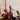 The Hall of Flame Jam Purple Wax Candle Holder, crafted from sustainable plant-based wax, stands lit among brown and yellow flowers, fresh figs, and glass vases on a light surface against a neutral background.