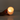 A hand holds the Hall of Flame Pillar with Wax Sleeve, its gentle light glowing warmly in a round holder against a plain backdrop, in compliance with open flame policy.