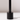 A close-up of the Hall of Flame Black Wax Candle Holder, featuring a minimalistic design with a vertical rod, casts a soft shadow on a white surface and background.