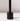 A close-up of the Hall of Flame Black Wax Candle Holder, featuring a minimalistic design with a vertical rod, casts a soft shadow on a white surface and background.
