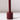 The Hall of Flame Bordeaux Dark Red Wax Candle Holder, featuring a cylindrical design with a central vertical rod and built-in safety mechanism, casts a shadow on a glossy white surface against a light background.
