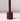 The Hall of Flame Bordeaux Dark Red Wax Candle Holder, featuring a cylindrical design with a central vertical rod and built-in safety mechanism, casts a shadow on a glossy white surface against a light background.