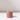 The Hall of Flame Dusty Rose Wax Candle Holder, with its pink cylindrical design and center hole, sits on a white reflective surface—ideal for showcasing your favorite pillar candle in stylish simplicity.