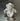 The Hall of Flame Mini Flower Object, a white ruffled decorative sculpture with a pearly sheen, rests on a reflective metallic surface, casting a gentle shadow below.