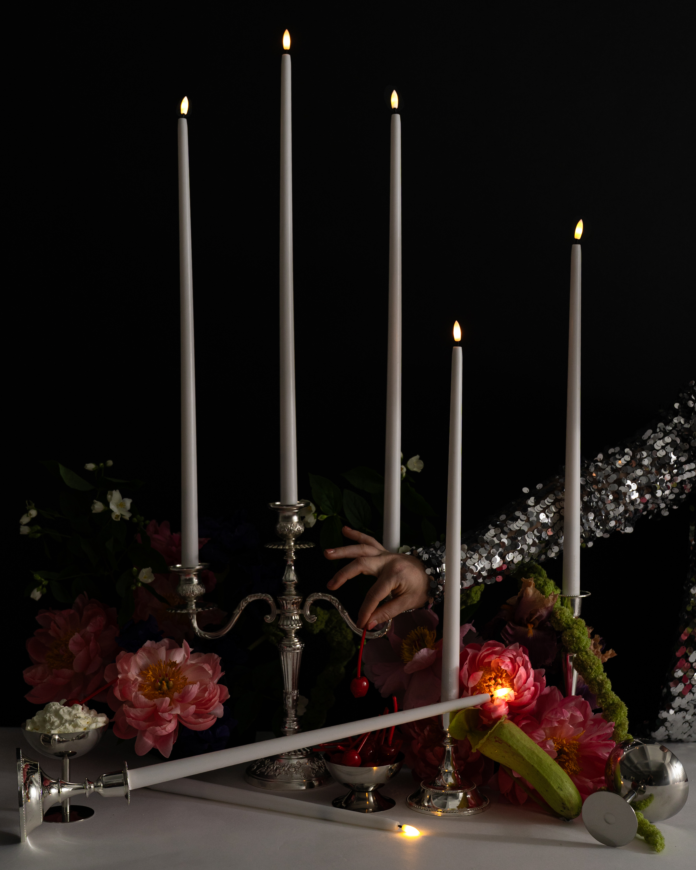 A hand in a sequined sleeve reaches for a silver five-candle candelabrum with Hall of Flame ULTRABRIGHT™ LED Flameless Taper Candles among pink flowers, as one candle lies broken on a white table scattered with petals and small flames.
