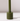 The Hall of Flame Moss Green Wax Candle Holder, a sustainable plant-based cylindrical piece with a tall, thin stem, stands on a white surface casting a faint rightward shadow against a softly lit plain background.