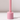 The Hall of Flame Petal Pink Wax Candle Holder features a cylindrical, glossy design with smooth edges. It stands on a white surface with a vertical rod, casting a soft shadow and offering minimalist elegance.