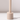 The Hall of Flame Sand Beige Wax Candle Holder, featuring a cylindrical shape and long vertical handle, stands upright on a white surface, casting a shadow to the right.