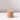 The Hall of Flame Venti Brown Wax Candle Holder, a cylindrical and beige sustainable piece with a central hole, rests on a white surface, casting a rightward shadow against a softly lit plain background.