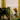 A pillar candle burns before napa cabbage, with cucumbers, bitter melon, and a yellow orchid in a glass vase. Hall of Flames Olive Green Wax Candle Holder and tall yellow cylinders are visible in the background.