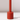 The Hall of Flame Poppy Red Wax Candle Holder, a tall cylindrical piece, stands upright on a white surface casting a small shadow to the right. Its plain light background highlights its sustainable plant-based wax design.