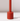 The Hall of Flame Poppy Red Wax Candle Holder, a tall cylindrical piece, stands upright on a white surface casting a small shadow to the right. Its plain light background highlights its sustainable plant-based wax design.