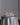 A Hall of Flame Round Table stands out in the room, elegantly covered with a silver satin cloth, gold flatware, two wine glasses, white flowers centerpiece, and Pillar Cluster candles from Hall of Flame, set against a plain gray wall.