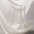 Two tall white candles are lit in Hall of Flame White Flower Candle Holders, placed on silky white fabric for a serene and elegant look.