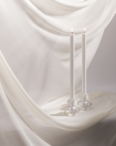 Two tall white candles are lit in Hall of Flame White Flower Candle Holders, placed on silky white fabric for a serene and elegant look.