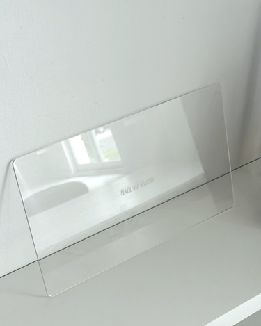 A clear, rectangular Acrylic Candle Drip Tray by Hall of Flame is displayed on a white shelf against a white wall, with the brand name faintly visible and a window reflection on the tray.