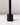 A close-up of the Hall of Flame Black Wax Candle Holder, featuring a minimalistic design with a vertical rod, casts a soft shadow on a white surface and background.