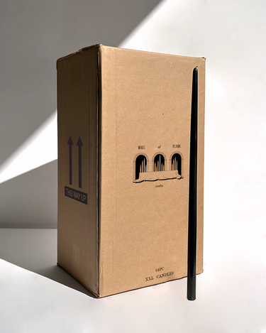 A tall, rectangular Hall of Flame Bulk Candles | Wholesale by-the-box packaging with THIS WAY UP arrows stands upright as a long black taper wedding candle leans against it, casting a shadow on the light background.