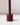 The Hall of Flame Bordeaux Dark Red Wax Candle Holder, featuring a cylindrical design with a central vertical rod and built-in safety mechanism, casts a shadow on a glossy white surface against a light background.
