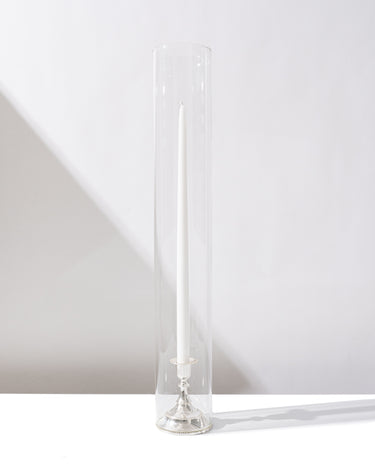 The Hall of Flame Short taper candle, set in an antique silver alloy holder and enclosed in a clear glass cylinder, stands upright on a white surface with its shadow in the background.