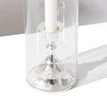 The Hall of Flame Short | Antique Silver Holder, crafted from alloy metal, features a white candle inside a tall, clear glass hurricane lamp and casts a shadow to the right on a white surface.