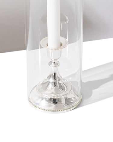 The Hall of Flame Short | Antique Silver Holder, crafted from alloy metal, features a white candle inside a tall, clear glass hurricane lamp and casts a shadow to the right on a white surface.