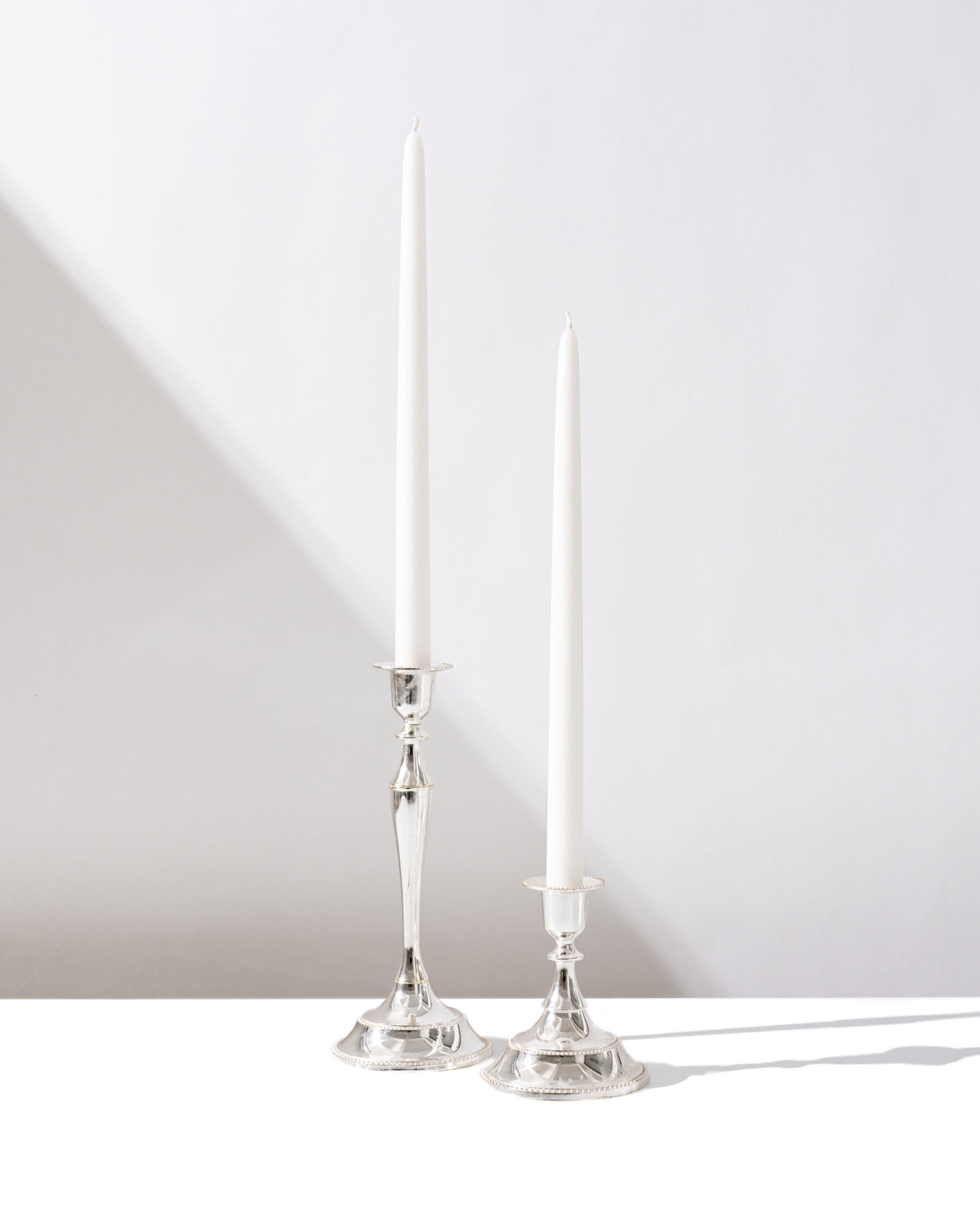 Two tall, white taper candles in Hall of Flames Antique Holder Duo | Silver cast soft shadows on a white surface. One antique silver candlestick is taller and more ornate than the other, both set against a light background.