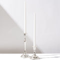 Two tall, white taper candles in Hall of Flames Antique Holder Duo | Silver cast soft shadows on a white surface. One antique silver candlestick is taller and more ornate than the other, both set against a light background.