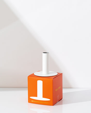 The Hall of Flame White Plated Candle Holder sits on an orange box with white text, ideal for displaying XXL Tapers, placed on a white surface against a light backdrop with a subtle shadow.