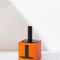 The Hall of Flame Black Plated Candle Holder, designed for XXL Tapers, rests atop an orange box with a matching black silhouette. The scene is brightly lit with distinct shadows on a white background.