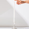 A hand reaches toward the unlit wick of a Hall of Flame White | Realistic LED Flameless Taper Candle, which stands upright in a shiny silver holder against a plain white background.