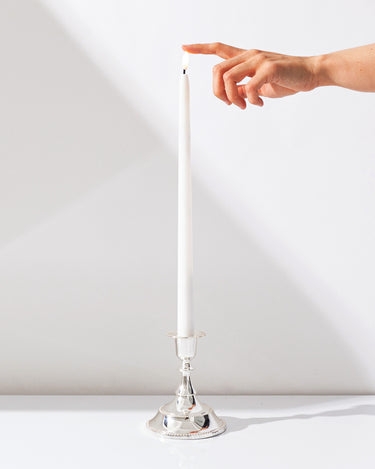 A hand reaches toward the unlit wick of a Hall of Flame White | Realistic LED Flameless Taper Candle, which stands upright in a shiny silver holder against a plain white background.
