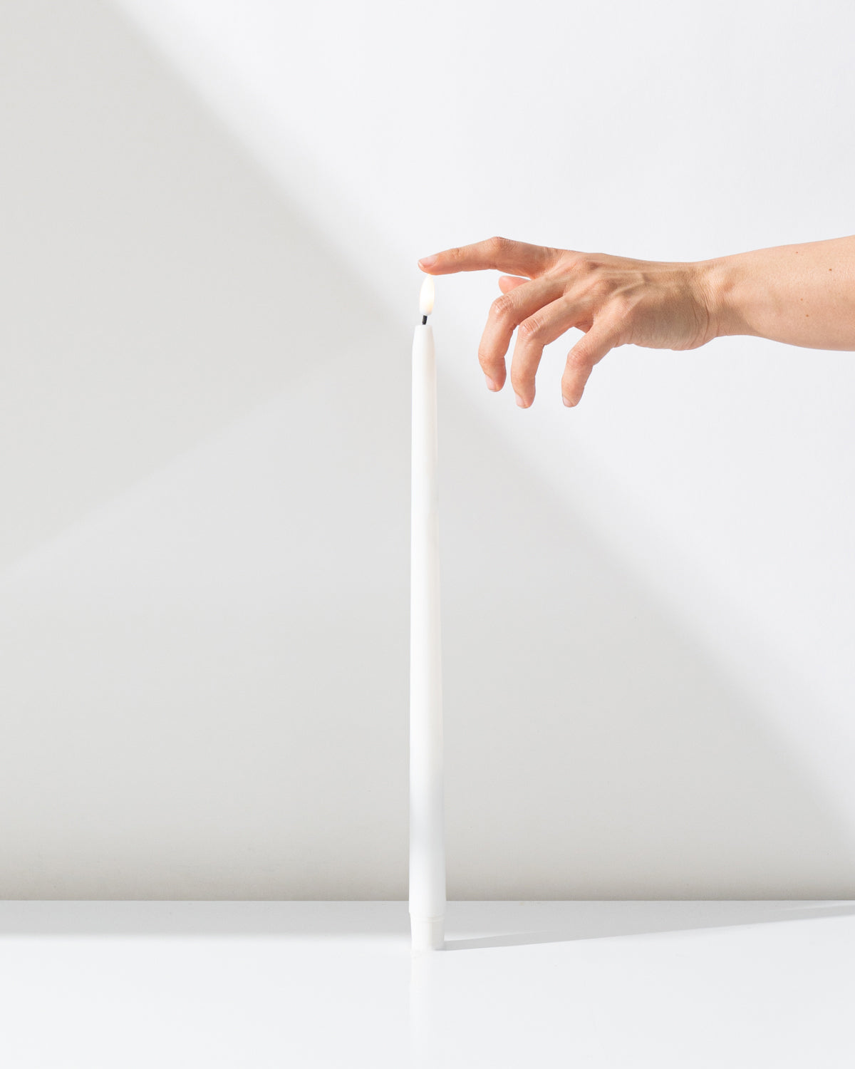 A hand gently touches the top of a Hall of Flame White Realistic LED Flameless Taper Candle, standing upright on a white surface against a white background.