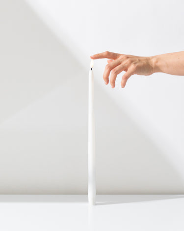 A hand gently touches the top of a Hall of Flame White Realistic LED Flameless Taper Candle, standing upright on a white surface against a white background.