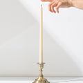 A hand reaches for the flame of a Hall of Flame Ivory Ultra-Bright LED Flameless Taper Candle, displayed in a gold holder on a white surface against a plain white background.
