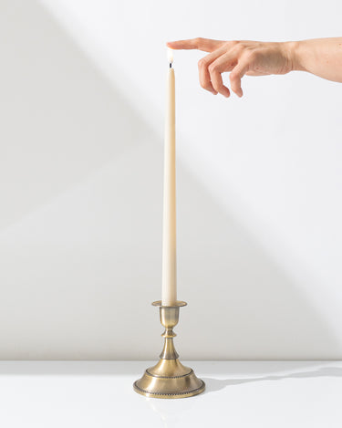 A hand reaches for the flame of a Hall of Flame Ivory Ultra-Bright LED Flameless Taper Candle, displayed in a gold holder on a white surface against a plain white background.
