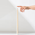 A hand reaches toward the soft glow of a Hall of Flame Ivory Ultra-Bright LED Flameless Taper Candle—a tall, slender beige candle standing upright on a white surface against a white background.