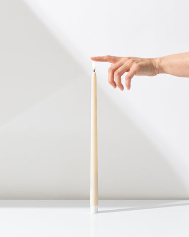A hand reaches toward the soft glow of a Hall of Flame Ivory Ultra-Bright LED Flameless Taper Candle—a tall, slender beige candle standing upright on a white surface against a white background.