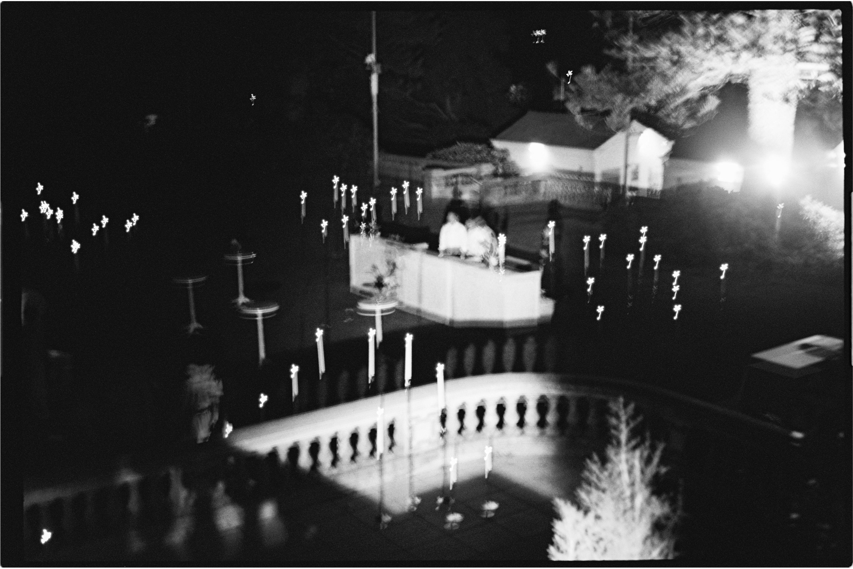 A mysterious, dimly lit black-and-white night scene shows two people at a table surrounded by Hall of Flame 8-in-1 Convertible Candle Holder lights on poles, with trees and a building softly outlined in the background.