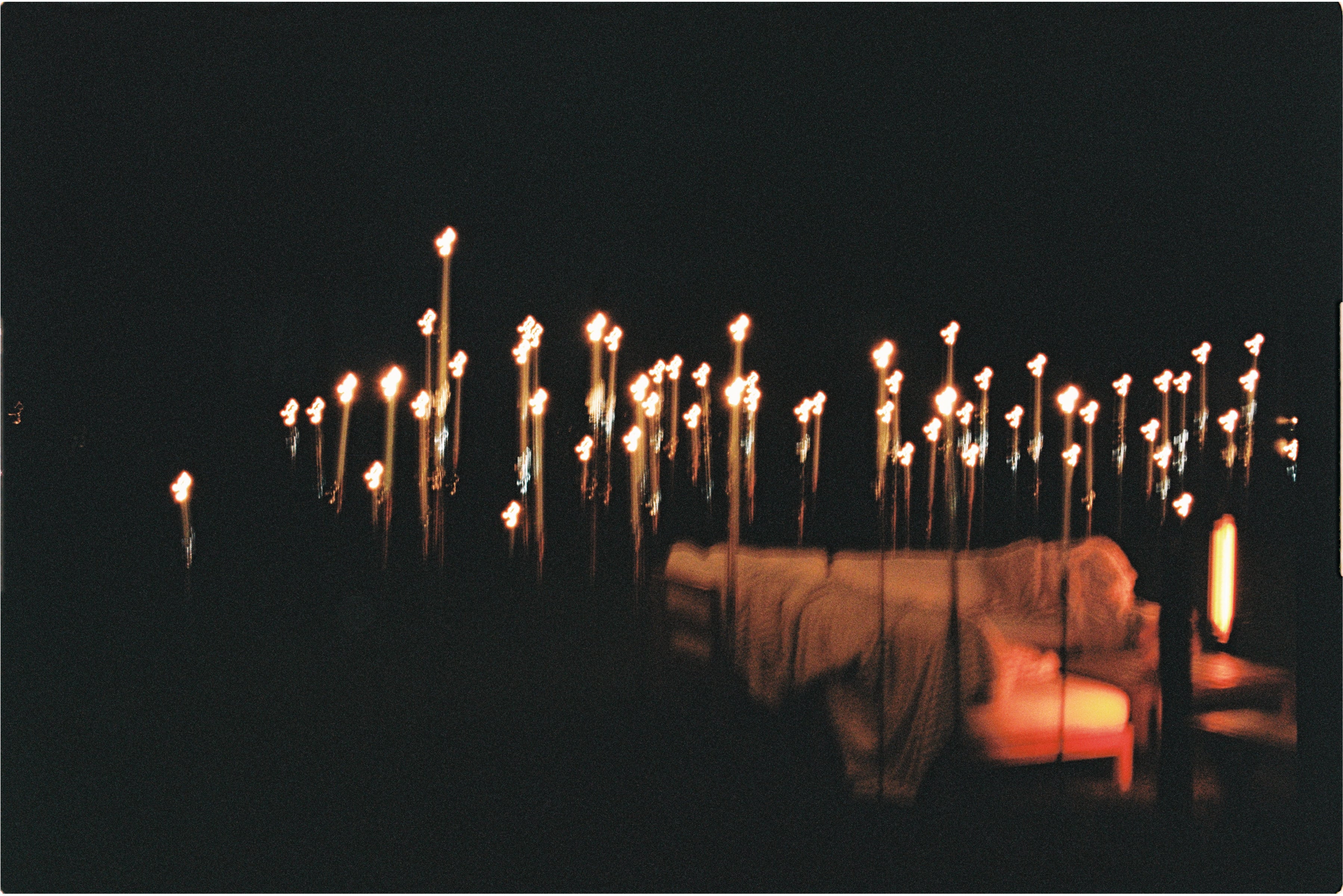 A dreamy, ethereal scene features a bed bathed in orange light, surrounded by Hall of Flame's 8-in-1 Convertible Candle Holder with ground stake and floor bases, glowing against a dark backdrop.
