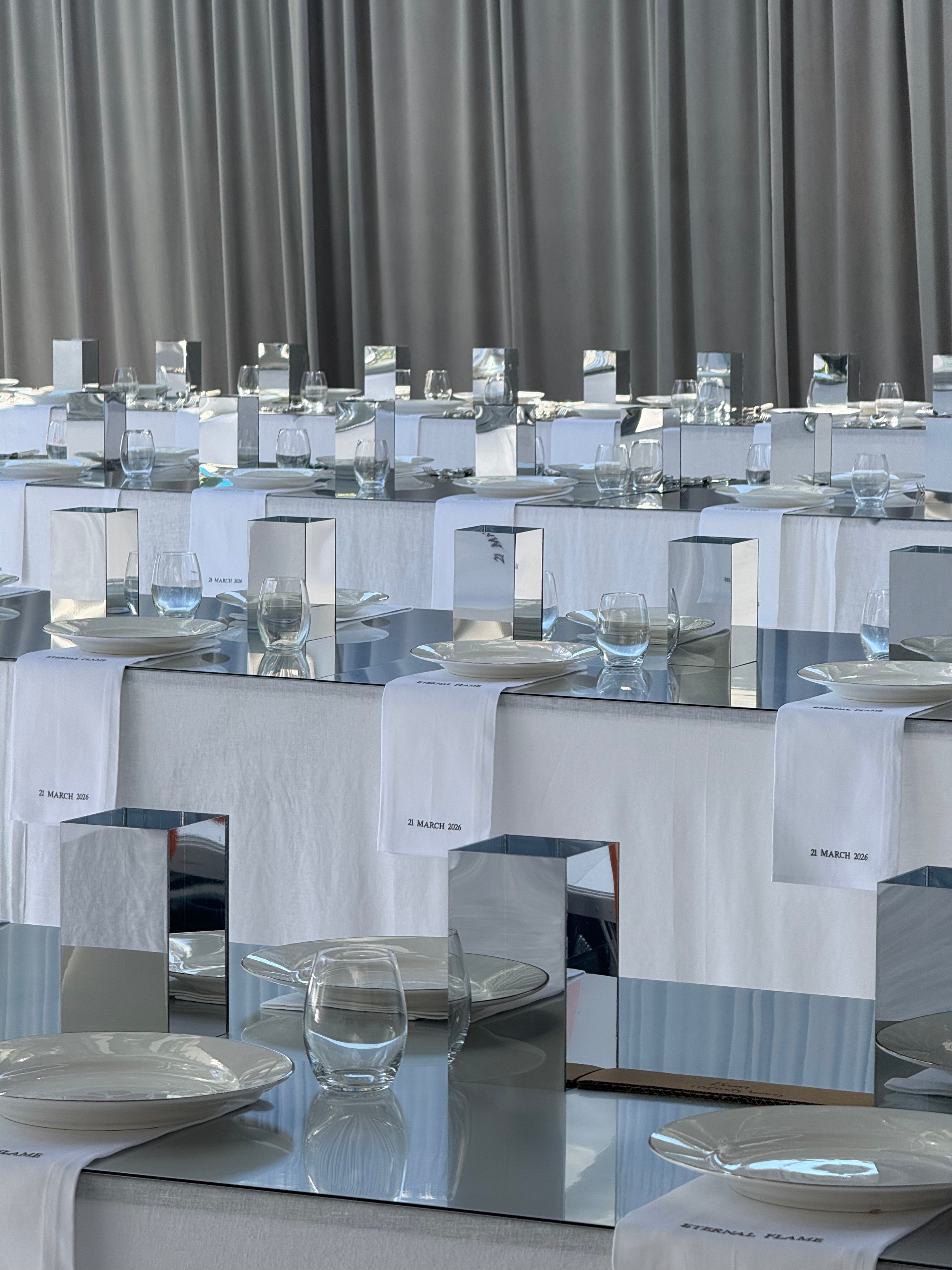 Tables set with white tablecloths, plates, glasses, and rectangular mirrors, with white cloth napkins and silver accents, in front of gray curtains.