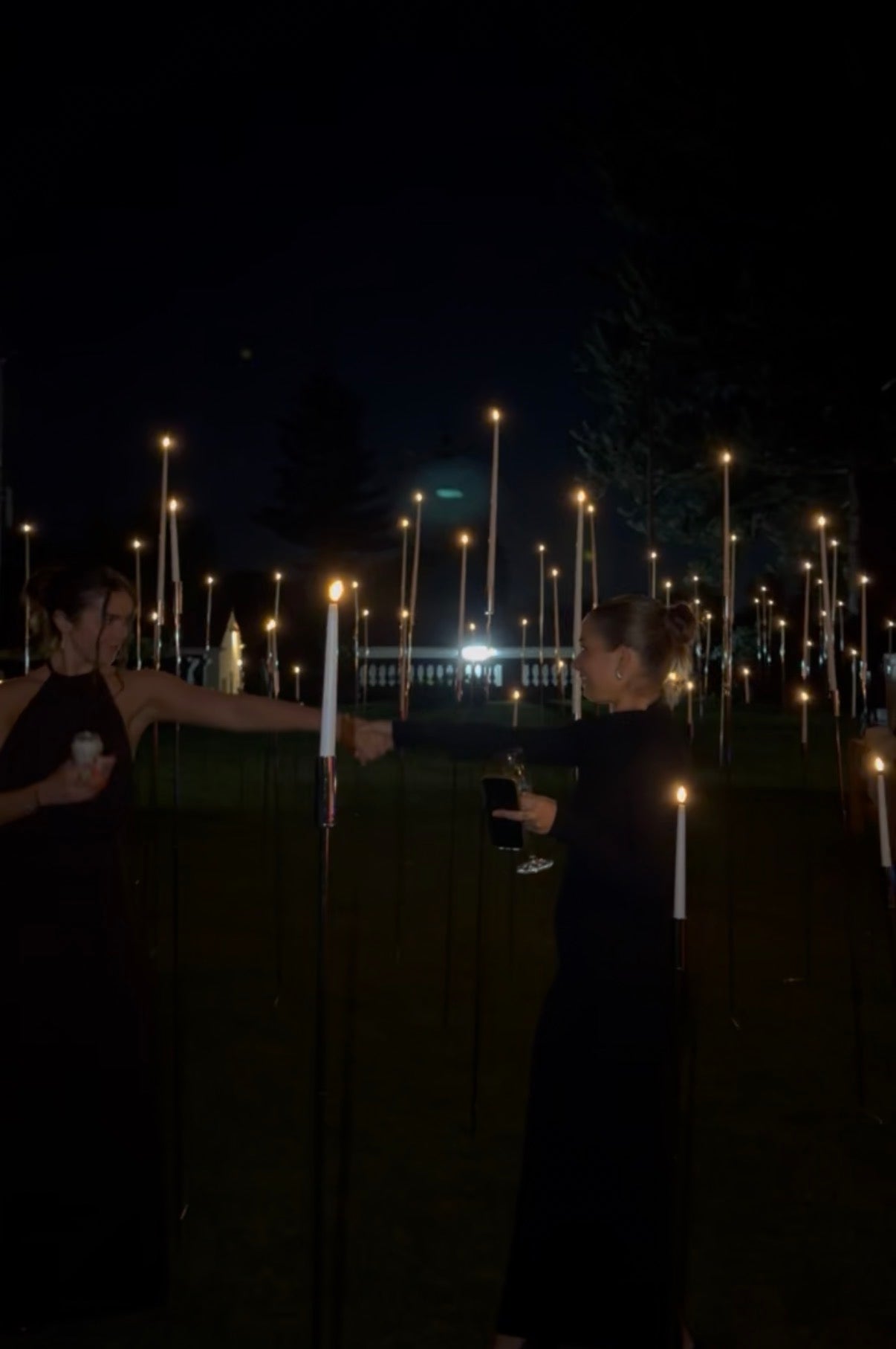 Two women in dark dresses stand outside at night, smiling and fist-bumping with drinks in hand, surrounded by glowing Hall of Flame 8-in-1 Convertible Candle Holder - Ground Stake & Floor Bases illuminating the trees.