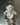The Hall of Flame Mini Flower Object, a white ruffled decorative sculpture with a pearly sheen, rests on a reflective metallic surface, casting a gentle shadow below.