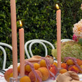 A table set outdoors features peach-colored candles, fresh peaches, pink ribbon, beige napkins, and flowers, with white chairs and green bushes in the background.