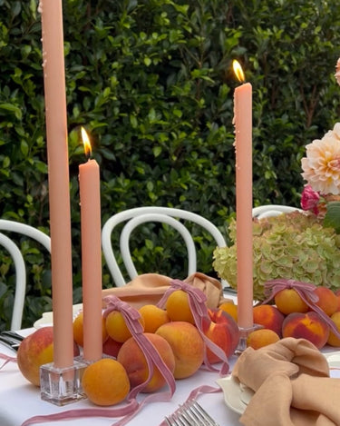 A table set outdoors features peach-colored candles, fresh peaches, pink ribbon, beige napkins, and flowers, with white chairs and green bushes in the background.