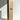 A tall, plain box labeled “Bulk Candles | Wholesale by-the-box” from Hall of Flame stands upright on a white surface, with a long, cream-colored candle leaning beside it—ideal for weddings or anyone seeking bulk candles. Soft sunlight casts gentle shadows.