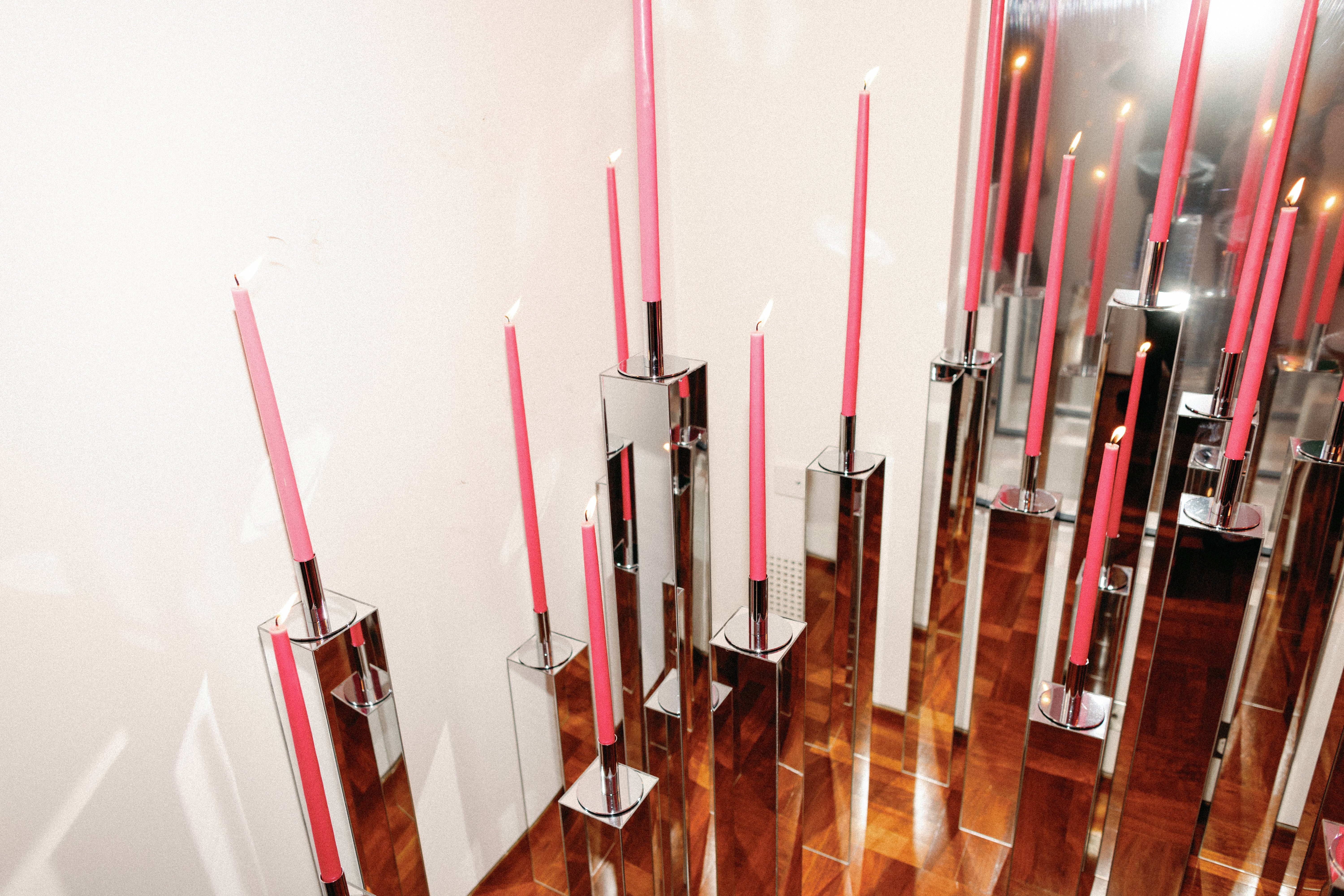 Several tall, pink candles are lit and placed in modern, mirrored candle holders of varying heights on a polished wooden floor near a white wall. The candle holders reflect the candles and surrounding light.